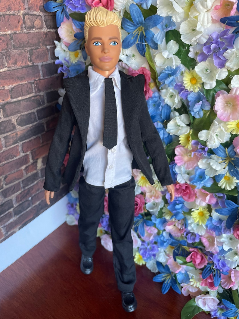 Puede incluir: Una mu&ntilde;eca que lleva un traje negro con una camisa blanca y una corbata negra. La mu&ntilde;eca tiene el pelo rubio y los ojos azules. La mu&ntilde;eca est&aacute; de pie frente a una pared de flores artificiales.