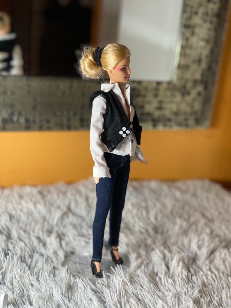 May include: A blonde doll wearing a white button-down shirt, a black vest with white buttons, dark blue jeans, and black heels.