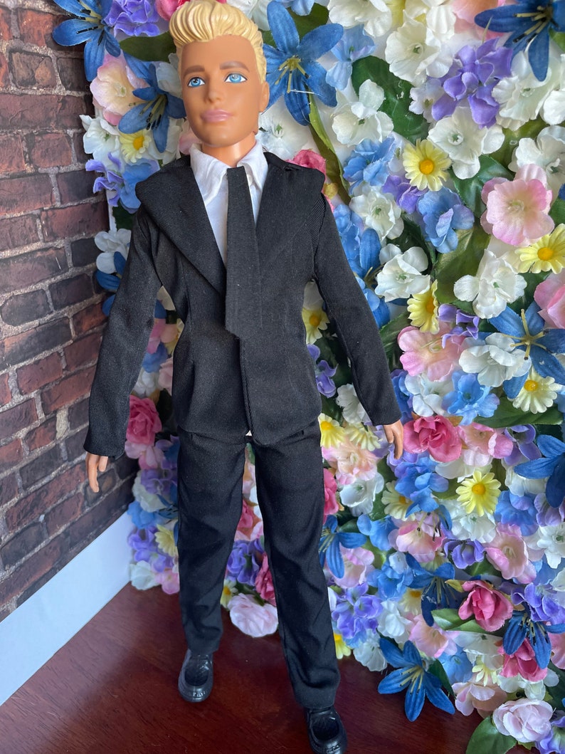 Puede incluir: Un traje negro con camisa blanca y corbata negra. El traje lo lleva una mu&ntilde;eca con el pelo rubio y los ojos azules. La mu&ntilde;eca est&aacute; de pie frente a una pared de flores artificiales.