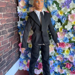 Puede incluir: Una mu&ntilde;eca vestida con un traje negro con camisa blanca y corbata negra. La mu&ntilde;eca tiene el pelo rubio y los ojos azules. La mu&ntilde;eca est&aacute; de pie frente a una pared de flores artificiales.