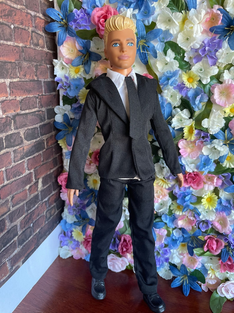 Puede incluir: Una mu&ntilde;eca vestida con un traje negro con camisa blanca y corbata negra. La mu&ntilde;eca tiene el pelo rubio y los ojos azules. La mu&ntilde;eca est&aacute; de pie frente a una pared de flores artificiales.