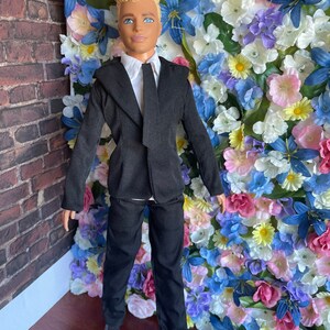 Puede incluir: Una mu&ntilde;eca vestida con un traje negro con camisa blanca y corbata negra. La mu&ntilde;eca tiene el pelo rubio y los ojos azules. La mu&ntilde;eca est&aacute; de pie frente a una pared de flores artificiales.