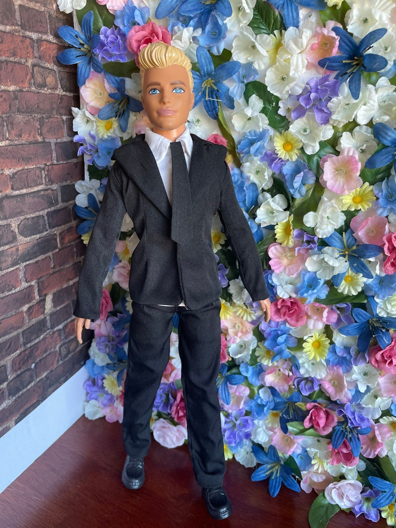 Puede incluir: Una mu&ntilde;eca vestida con un traje negro con camisa blanca y corbata negra. La mu&ntilde;eca tiene el pelo rubio y los ojos azules. La mu&ntilde;eca est&aacute; de pie frente a una pared de flores artificiales.
