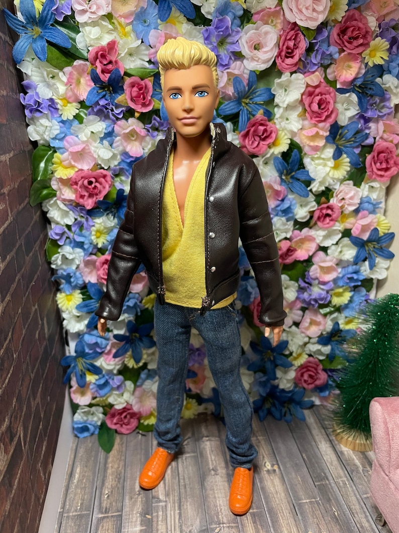 May include: A doll wearing a brown leather jacket, a yellow sweater, blue jeans, and orange shoes. The doll is standing in front of a wall of artificial flowers.