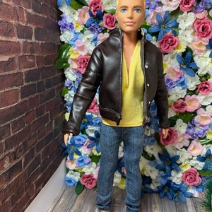May include: A doll wearing a brown leather jacket, a yellow shirt, blue jeans, and orange shoes. The doll is standing in front of a wall of artificial flowers.