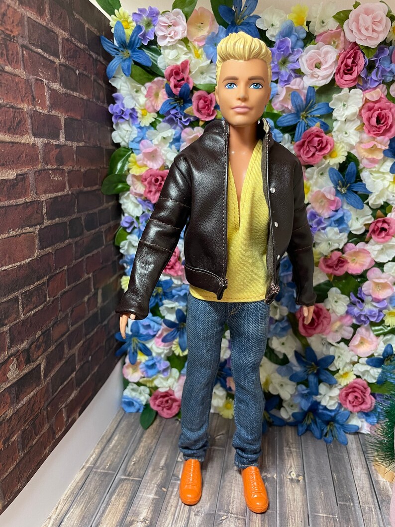 May include: A doll wearing a brown leather jacket, a yellow shirt, blue jeans, and orange shoes. The doll is standing in front of a wall of artificial flowers.