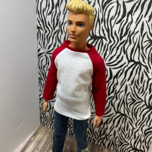 May include: A doll wearing a white and red baseball shirt, blue jeans, and orange shoes. The doll has blonde hair and blue eyes.