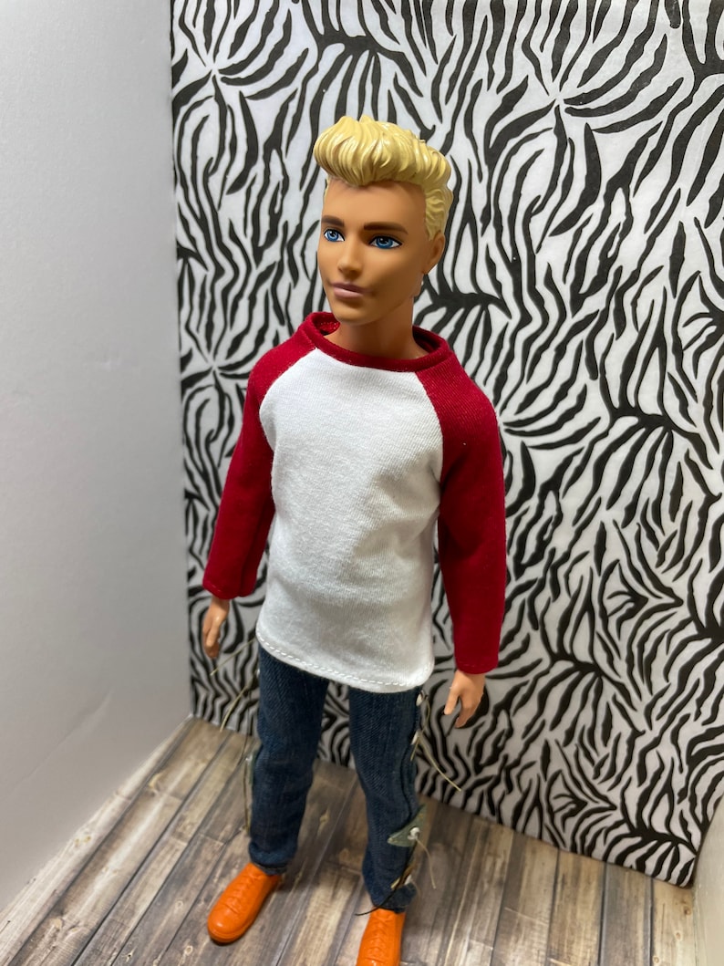 May include: A doll wearing a white and red baseball shirt, blue jeans, and orange shoes. The doll has blonde hair and blue eyes.