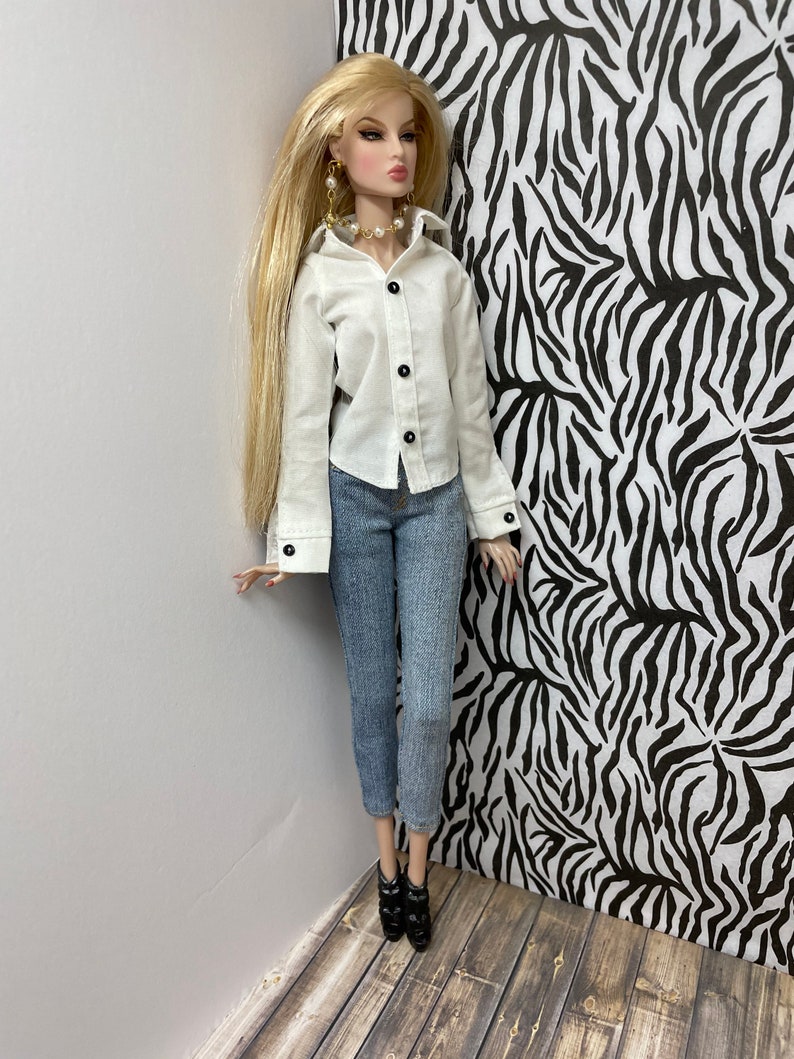 May include: A doll wearing a white button-down shirt with black buttons, blue jeans, and black heeled ankle boots. The doll has long blonde hair and is wearing pearl earrings and a pearl necklace.