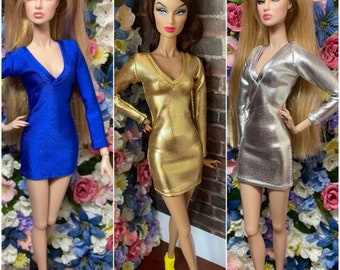 Puede incluir: Tres muñecas de moda con minivestidos de manga larga. Los vestidos son azules, dorados y plateados. Las muñecas tienen el pelo rubio y castaño. La muñeca del centro lleva zapatos amarillos. Las muñecas posan frente a un fondo floral.