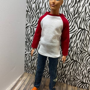 May include: A doll wearing a white and red baseball shirt, blue jeans with silver accents, and orange shoes.