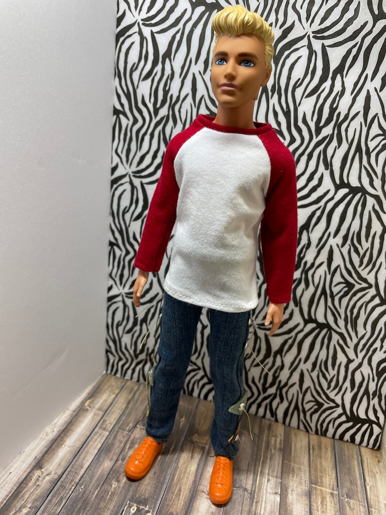 May include: A doll wearing a white and red baseball shirt, blue jeans with silver accents, and orange shoes.