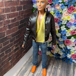 May include: A fashion doll wearing a brown leather jacket, a yellow shirt, blue jeans, and orange shoes. The doll is standing in front of a brick wall and a wall of flowers.
