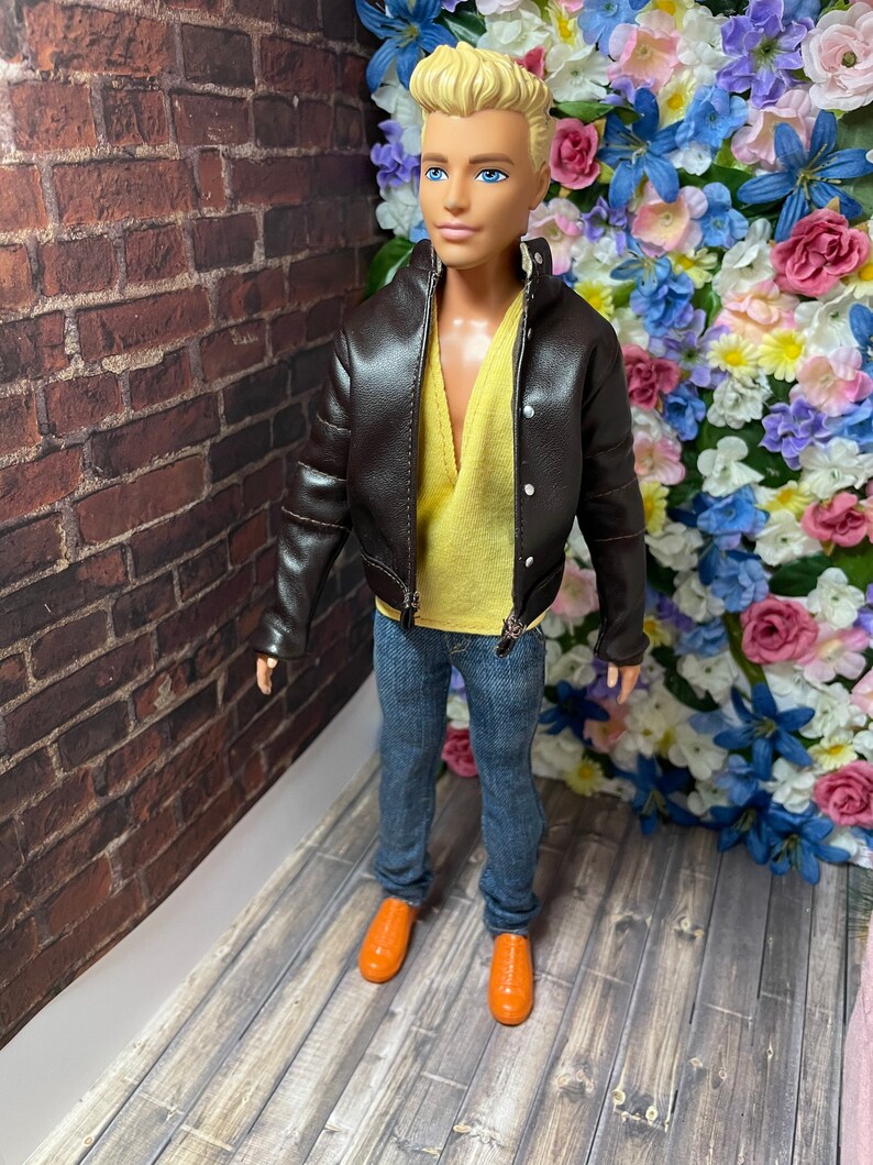 May include: A fashion doll wearing a brown leather jacket, a yellow shirt, blue jeans, and orange shoes. The doll is standing in front of a brick wall and a wall of flowers.