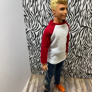 May include: A doll wearing a white and red baseball shirt, blue jeans, and orange shoes. The doll has blonde hair and blue eyes.