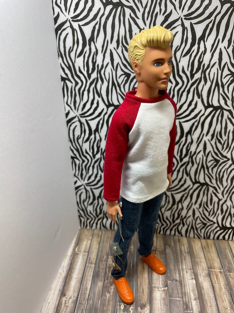 May include: A doll wearing a white and red baseball shirt, blue jeans, and orange shoes. The doll has blonde hair and blue eyes.