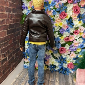 May include: A doll wearing a brown leather jacket, a yellow t-shirt, blue jeans, and brown shoes. The doll is standing in front of a wall covered in colourful artificial flowers.