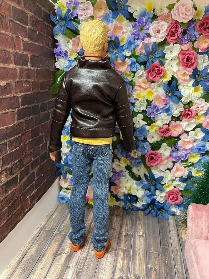 May include: A doll wearing a brown leather jacket, a yellow t-shirt, blue jeans, and brown shoes. The doll is standing in front of a wall covered in colourful artificial flowers.