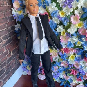 Puede incluir: Una mu&ntilde;eca vestida con un traje negro con camisa blanca y corbata negra. La mu&ntilde;eca tiene el pelo rubio y los ojos azules. La mu&ntilde;eca est&aacute; de pie frente a una pared de flores artificiales.