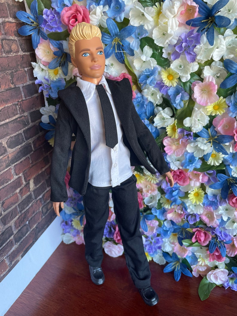 Puede incluir: Una mu&ntilde;eca vestida con un traje negro con camisa blanca y corbata negra. La mu&ntilde;eca tiene el pelo rubio y los ojos azules. La mu&ntilde;eca est&aacute; de pie frente a una pared de flores artificiales.