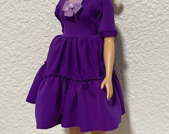 May include: A purple dress with a ruffled skirt and a lavender flower accent. The doll is wearing pink shoes.