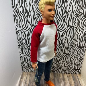May include: A doll wearing a white and red baseball shirt, blue jeans, and orange shoes. The doll has blonde hair and blue eyes.