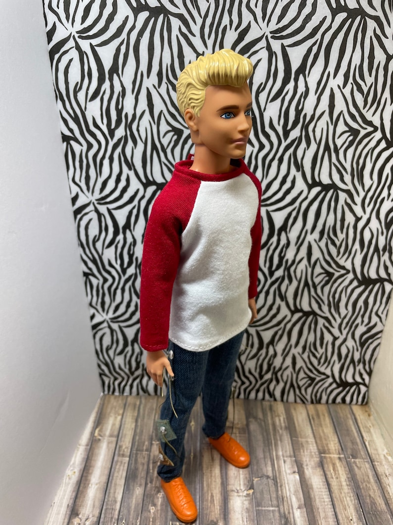 May include: A doll wearing a white and red baseball shirt, blue jeans, and orange shoes. The doll has blonde hair and blue eyes.