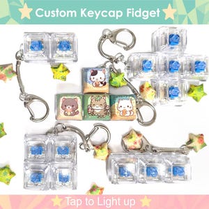 May include: Four clear keycap fidgets with light up blue buttons, each attached to a silver chain with a key ring. The keycaps feature different cartoon animal designs. The text "Tap to Light up" is printed below the keycaps.