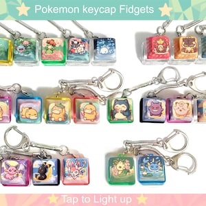 May include: A collection of square keycap fidget keychains featuring various colorful cartoon character illustrations. Each translucent plastic keycap is attached to a silver metal keyring. The image includes the text Pokemon keycap Fidgets and Tap to Light up.
