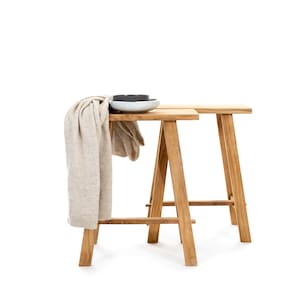 Puede incluir: Dos taburetes de madera con un acabado marrón claro. Los taburetes están hechos de madera maciza y tienen un diseño simple y rústico. Una manta beige está drapeada sobre los taburetes.