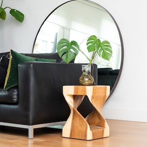 Könnte beinhalten: Ein modernes Wohnzimmer mit einem schwarzen Ledersofa, grünen und schwarzen Kissen und einem holzfarbenen Beistelltisch in Sanduhrform. Ein großer runder Spiegel und eine Vase mit Blättern sind ebenfalls zu sehen.