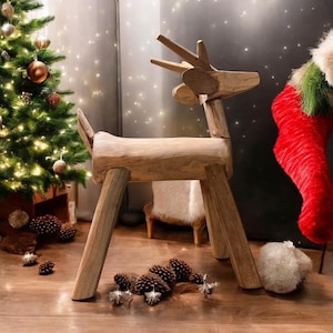 Puede incluir: Una escultura de reno de madera con un acabado marrón natural. El reno está de pie sobre un suelo de madera con piñas esparcidas a su alrededor. Un árbol de Navidad con luces y adornos está en el fondo.
