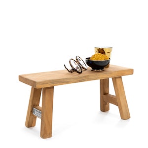 Peut inclure: Un petit banc en bois rectangulaire avec des pieds inclinés. Sur le banc se trouvent une paire de lunettes en écaille, un bol noir avec une bordure dorée rempli de graines jaunes et un verre doré. Le banc a une finition en grain de bois naturel.