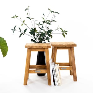 Op de afbeelding: Twee houten krukken met een zwarte vaas en een stapel boeken op een van de krukken. De krukken zijn gemaakt van natuurlijk hout en hebben een eenvoudig ontwerp.