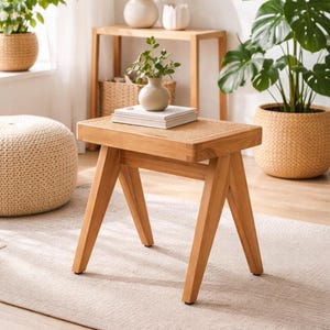 Puede incluir: Un taburete de madera marrón claro con una parte superior tejida. El taburete tiene patas anguladas y está colocado sobre una alfombra de color claro. Plantas en cestas tejidas y una mesa pequeña con una lámpara están en el fondo.
