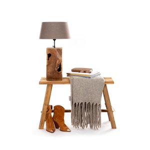 Op de afbeelding: Een houten tafellamp met een bruine lampenkap staat op een kleine houten bank. Een beige franjedeken, boeken en bruine suède laarzen met goudkleurige metalen punten staan ook op de bank. De laarzen hebben een hak.