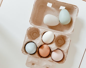May include: A cardboard egg carton with six pastel colored eggs. The eggs are in shades of blue, green, pink, and brown.