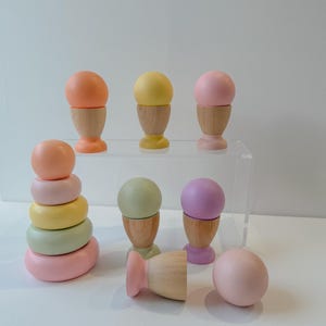 May include: A set of wooden stacking rings and egg cups in pastel colors. The rings are in shades of pink, green, and yellow, while the egg cups are in shades of pink, purple, and green. The egg cups are made of wood and have a simple design.