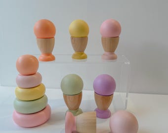 May include: A set of wooden stacking rings and egg cups in pastel colors. The rings are in shades of pink, green, and yellow, while the egg cups are in shades of pink, purple, and green. The egg cups are made of wood and have a simple design.