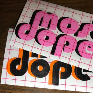 May include: Two sheets of white paper with a grid pattern. The top sheet has the words "most dope" in pink letters with black outlines. The bottom sheet has the words "the dope" in black letters with orange outlines.