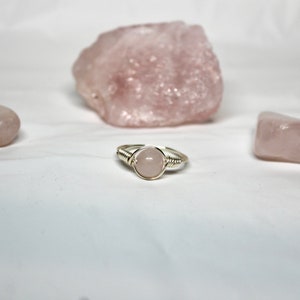 May include: A silver ring with a pink rose quartz stone wrapped in silver wire. The ring is surrounded by three pink rose quartz stones.