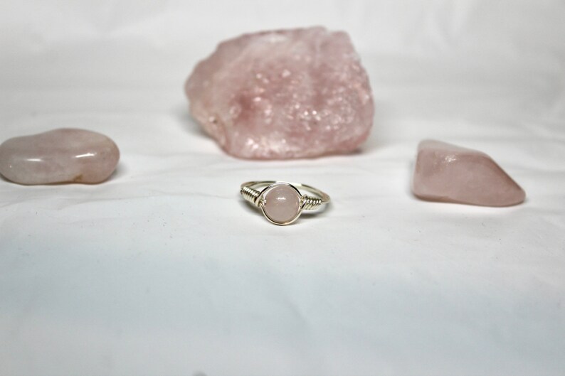 May include: A silver ring with a pink rose quartz stone wrapped in silver wire. The ring is surrounded by three pink rose quartz stones.