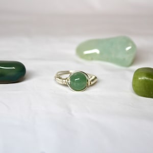 May include: A silver ring with a green gemstone set in the center. The ring is surrounded by silver wire. There are three green stones in the background.