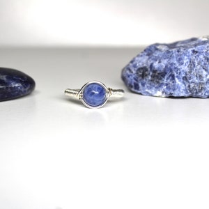 May include: A silver wire wrapped ring with a blue gemstone. The ring is sitting on a white surface next to two blue stones.