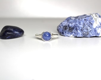 May include: A silver wire wrapped ring with a blue gemstone. The ring is sitting on a white surface next to two blue stones.