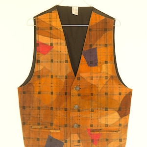 Op de afbeelding: Een bruin suède vest met een ruitpatroon en kleurrijke geometrische patches. Het vest heeft een V-hals en knoopsluiting.