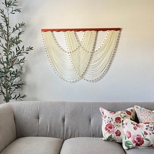 May include: A white macrame wall hanging with a wooden frame and black beads. The wall hanging is in the shape of two overlapping arches. The wall hanging is hanging on a white wall above a gray couch with floral patterned pillows.