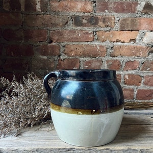 Puede incluir: Un tarro de barro vintage con un esmalte negro y una banda marrón y blanca alrededor de la parte superior. El tarro tiene una sola asa y está sentado sobre una superficie de madera.