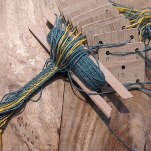 May include: A set of light-coloured wooden weaving cards with holes, threaded with teal and yellow yarn. The cards are arranged in a stack, with some labelled with the letters A, B, C and the number 10. The yarn extends from the cards in a long, braided tail.