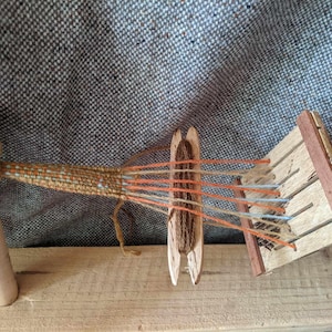 May include: A wooden weaving loom with orange, blue, and brown threads. The loom is set up on a wooden surface, with a woven piece in progress. The background is a textured grey fabric.